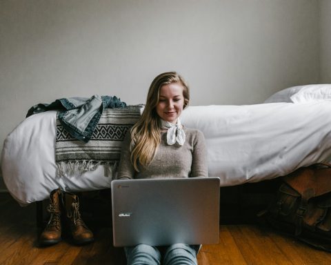 woman sitting beside a bed while using a laptop